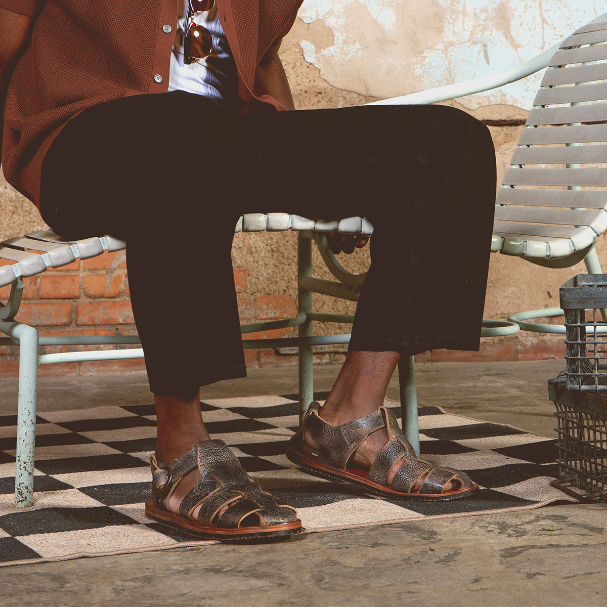 A person sits on a vintage metal chair, wearing black pants, a brown jacket, and Bed Stu's Lone strappy sandals featuring distressed leather straps and an antique brass buckle, with a tiled floor and weathered wall in the background.