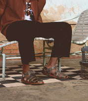 A person sits on a vintage metal chair, wearing black pants, a brown jacket, and Bed Stu's Lone strappy sandals featuring distressed leather straps and an antique brass buckle, with a tiled floor and weathered wall in the background.