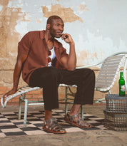 A man wears the Bed Stu Lone strappy sandals with an antique brass buckle, paired with a brown shirt and black pants as he sits on a white lounge chair. A green bottle and metal basket sit nearby on the checkered floor.