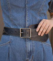 A person wearing a wide, dark Bed|Stü Lucen leather belt with a rectangular metal buckle over a denim dress.