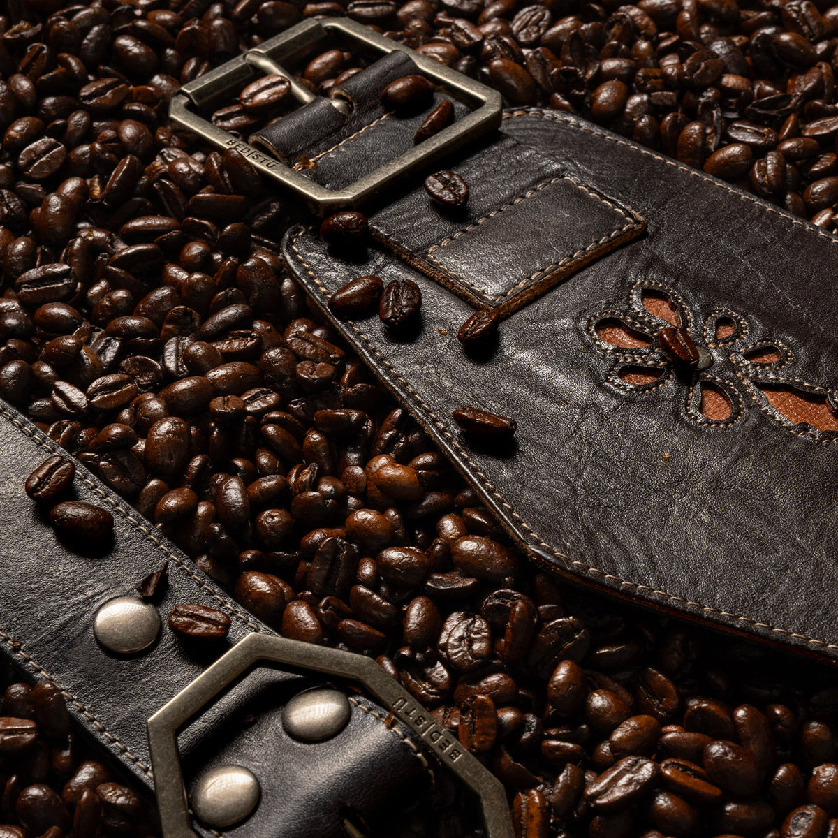 Bed|Stü Lucen dark brown leather boho-chic belt with a metal buckle and floral cutout detail, resting on a bed of roasted coffee beans.