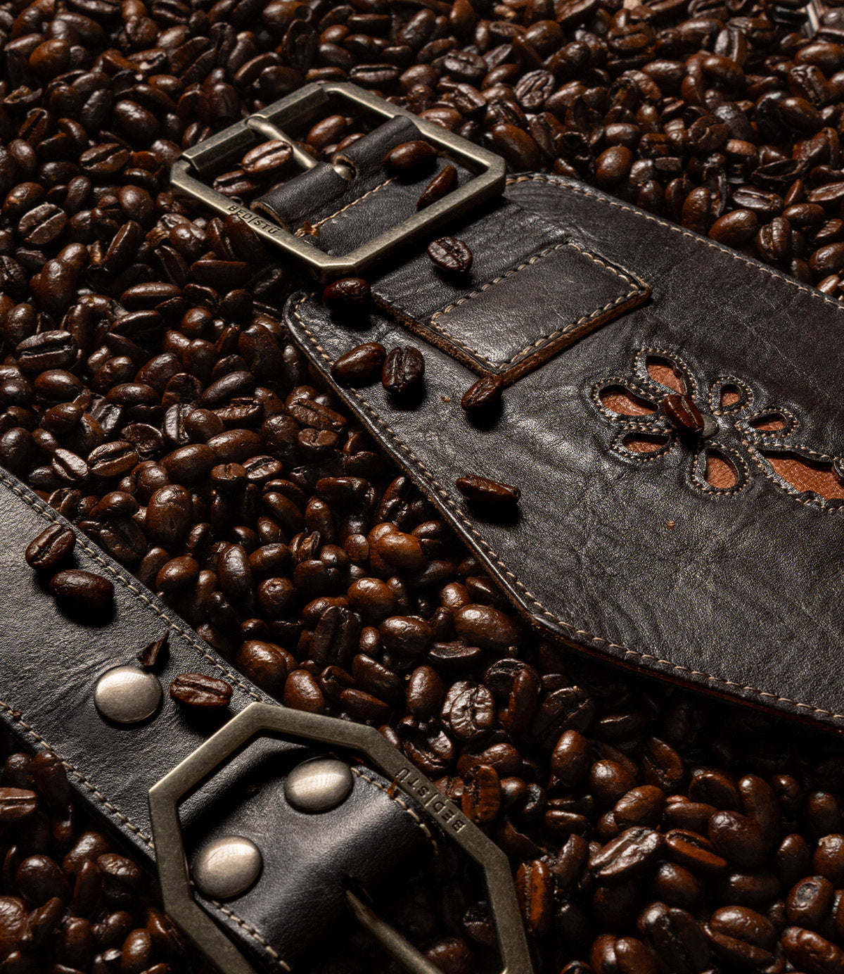 Bed|Stü Lucen dark brown leather boho-chic belt with a metal buckle and floral cutout detail, resting on a bed of roasted coffee beans.