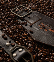 Bed|Stü Lucen dark brown leather boho-chic belt with a metal buckle and floral cutout detail, resting on a bed of roasted coffee beans.