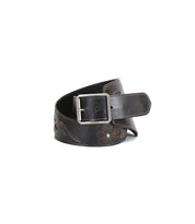 The Bed|Stü Lucen is a wide, dark leather boho-chic belt featuring a silver rectangular buckle and floral cutout patterns; shown coiled on a white background, this expressive accessory adds stylish flair to any ensemble.