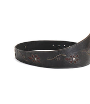 The Bed|Stü Lucen belt, featuring dark leather with boho-chic floral patterns and decorative stitching, is partially coiled on a white background and serves as a striking accessory that infuses any outfit with personality.