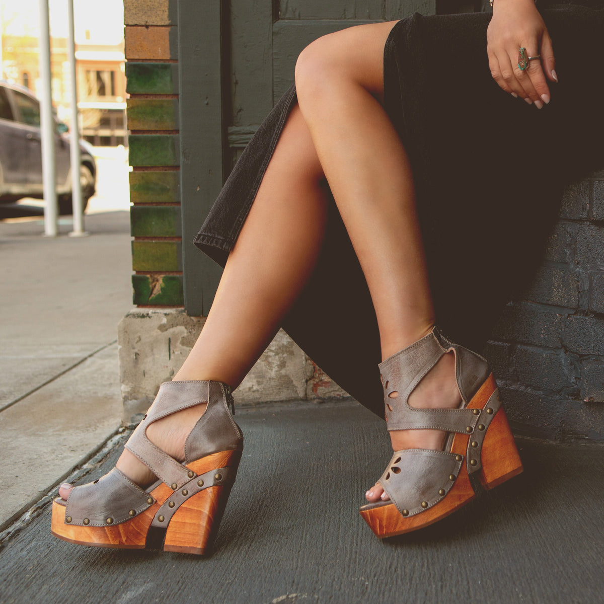 A person sitting on a sidewalk with their legs crossed, wearing Lucrative studded, wooden wedges from Bed Stu with zipper closure.