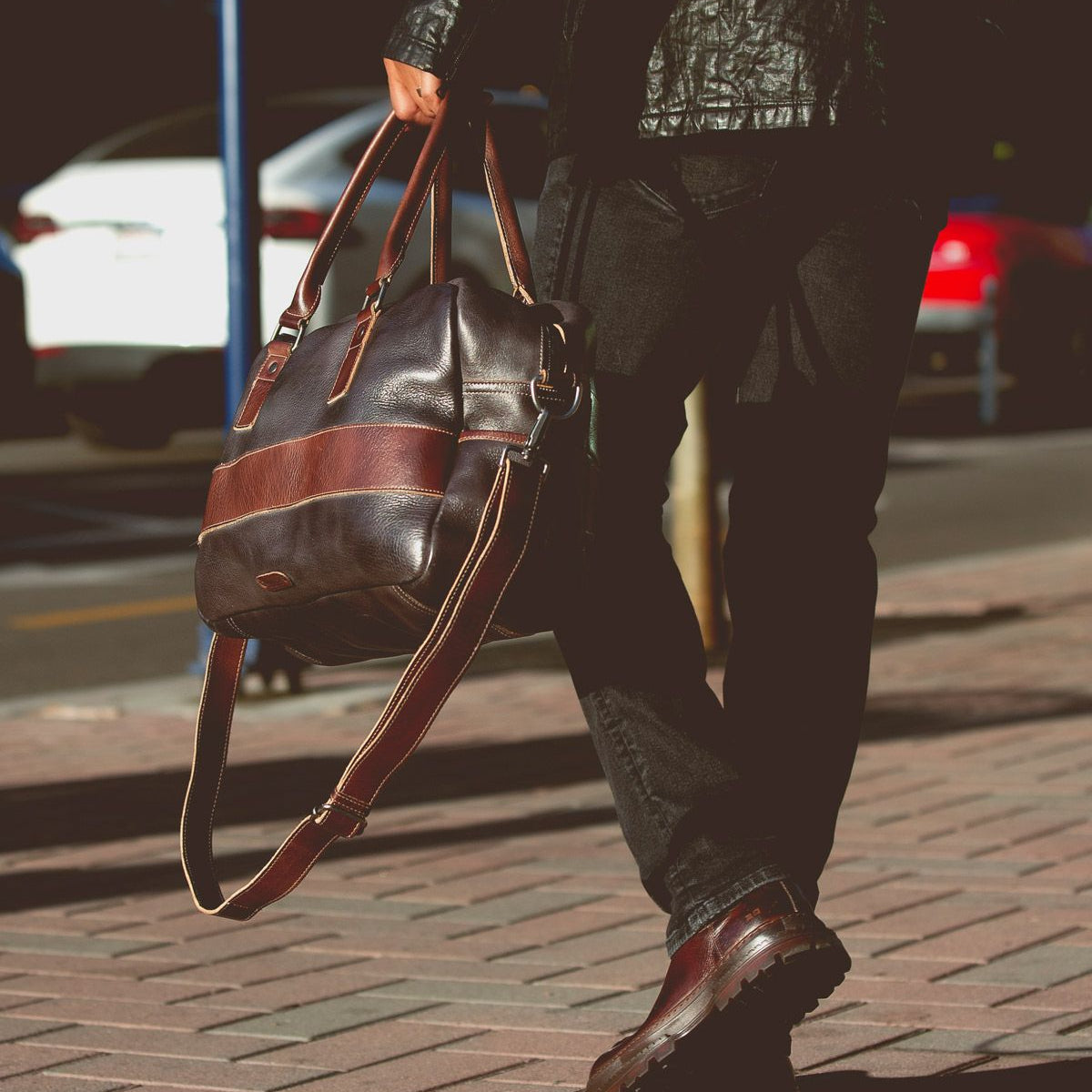 A person walking on a sidewalk is holding the Proem brown leather bag by Bed Stu, wearing a black leather jacket with a vintage metal belt buckle, black pants, and brown shoes.