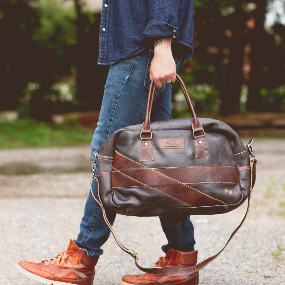 A person in blue jeans and a denim shirt walks while holding a stylish leather duffle in one hand and wearing brown high-top Luke sneakers from Bed Stu.