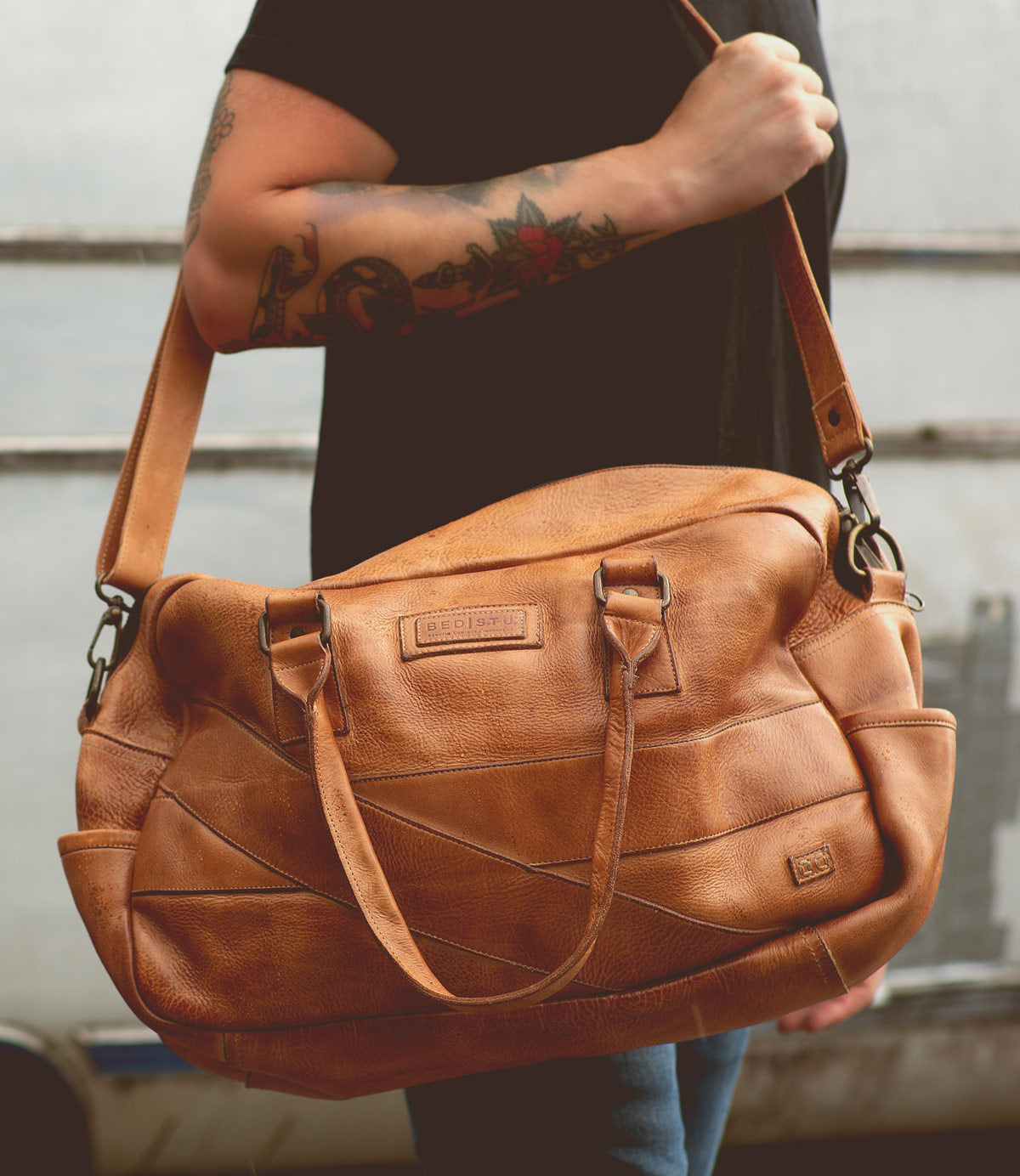 A person with tattooed arms holds a large, tan leather duffel bag by its adjustable crossbody strap and handles, the perfect travel companion: the Luke by Bed Stu.