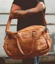 A person with tattooed arms holds a large, tan leather duffel bag by its adjustable crossbody strap and handles, the perfect travel companion: the Luke by Bed Stu.