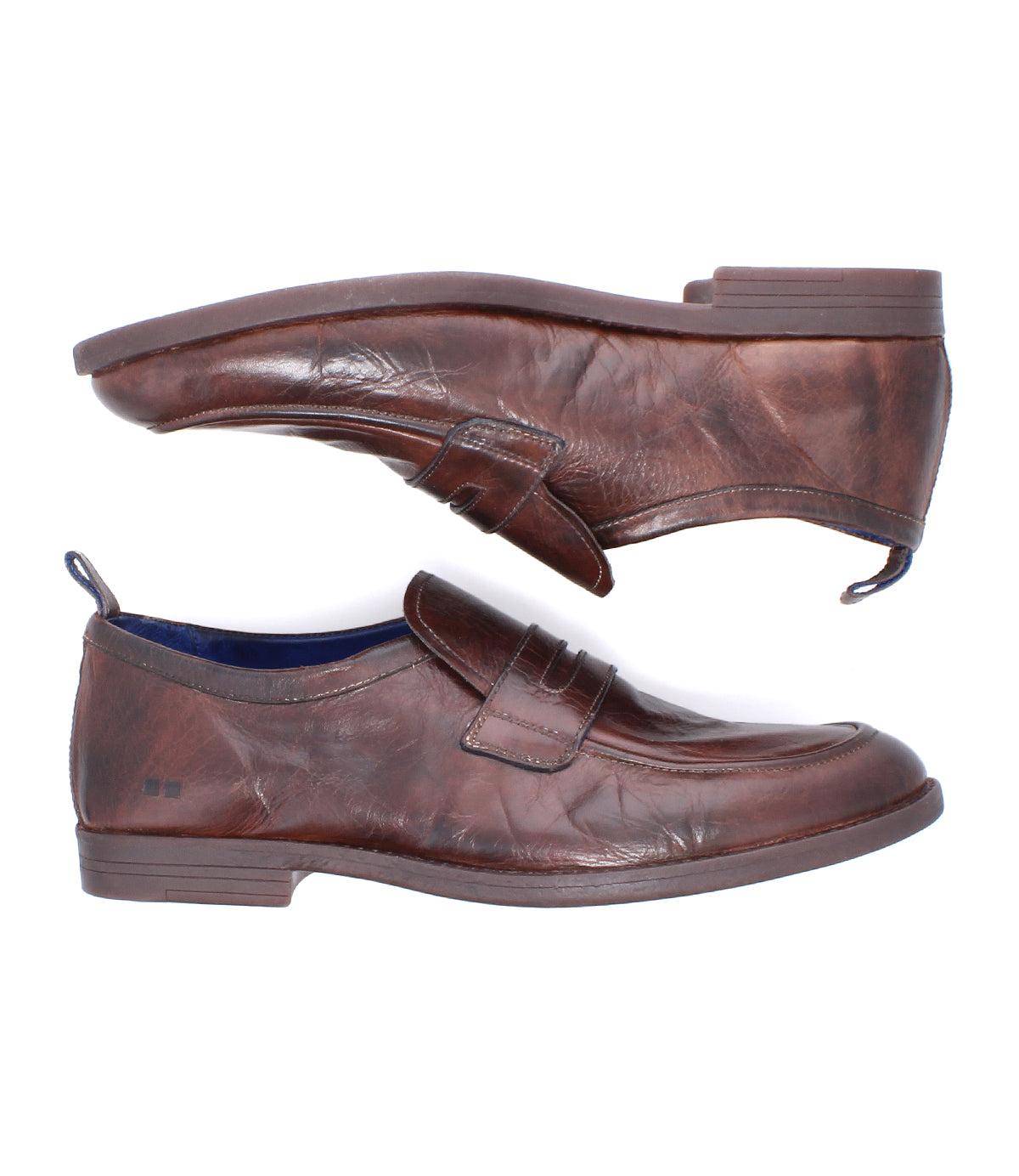 The Bed|Stü Lumaro features a pair of brown distressed leather loafers displayed from the side—one shoe facing right, the other above and facing left—where classic penny loafer style is infused with relaxed elegance.