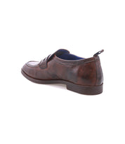 The Bed|Stü Lumaro is a brown leather penny loafer with a textured, distressed finish, low heel, and convenient pull tab at the back, presented at an angle on a white background—a seamless fusion of relaxed elegance and enduring style.