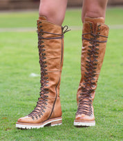 Close-up of a person wearing knee-high, brown leather Lustrous boots by Bed Stu with white soles and a cushioned insole, standing on a grassy surface.