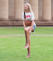 A person dressed casually in a white T-shirt, shorts, and Lustrous boots by Bed Stu with cushioned insoles stands on a grassy area in front of a building with tall columns.