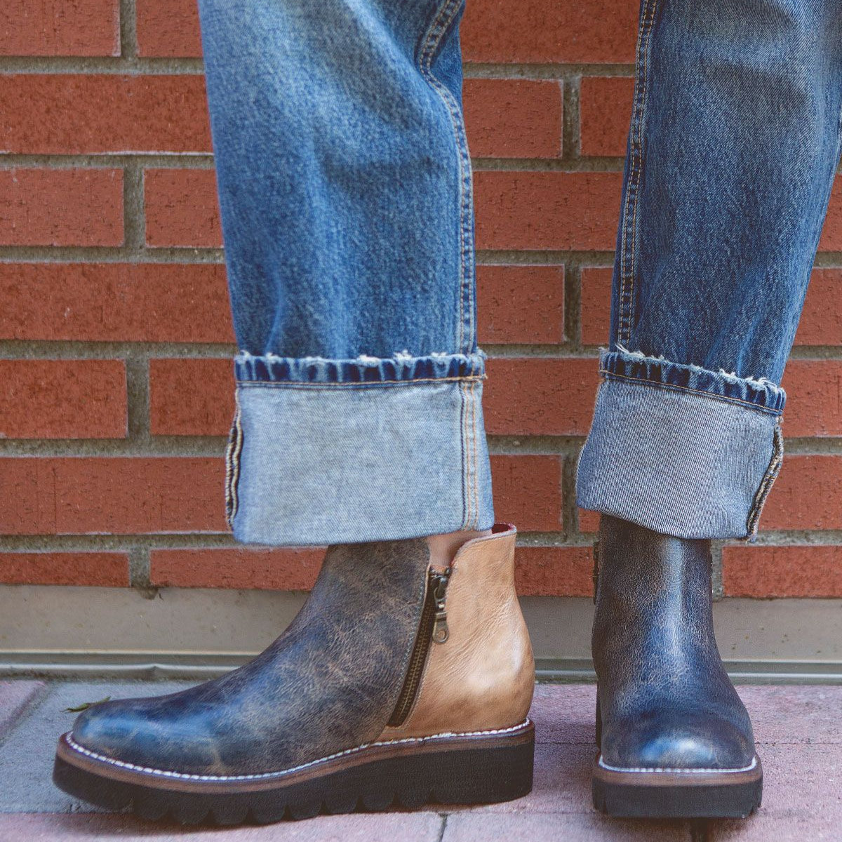 A person wearing cuffed jeans and a pair of Bed Stu's Lydyi two-tone leather ankle boots with a lightweight platform outsole stands on a brick-paved area near a brick wall.