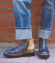 A person wearing cuffed jeans and a pair of Bed Stu's Lydyi two-tone leather ankle boots with a lightweight platform outsole stands on a brick-paved area near a brick wall.