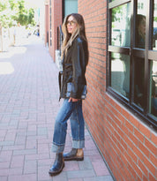 A woman wearing a black leather jacket, jeans, and Lydyi boots with a lightweight platform outsole by Bed Stu stands on a sidewalk next to a brick building, her reflection visible in the window.