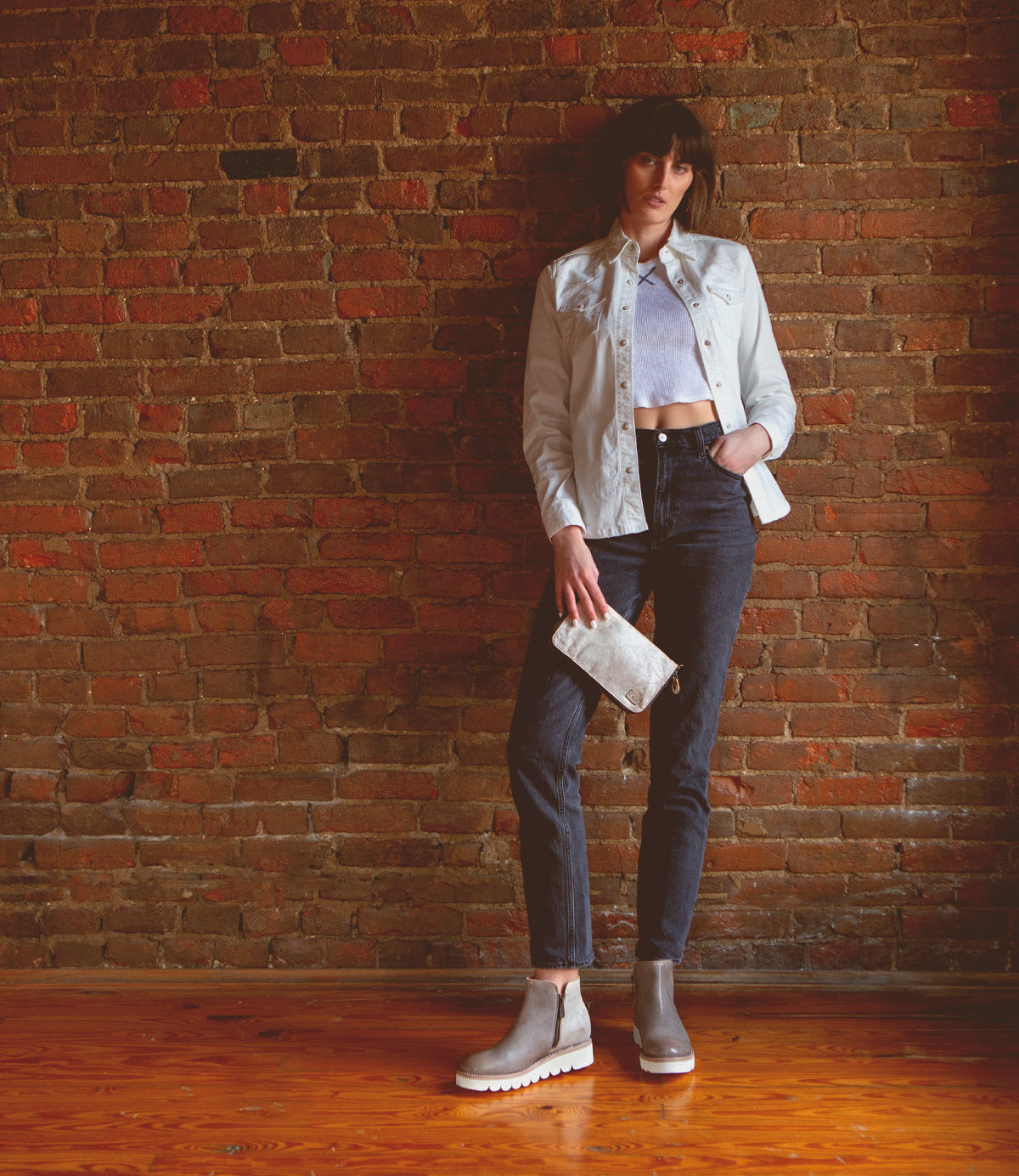 A woman leaning against a brick wall wearing jeans, a white jacket, and Bed Stu Lydyi leather ankle boots.