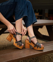 A person sitting on a wooden bench fastens the ankle strap of Bed Stu’s Lyxon open-toe, chunky high-heeled sandals, paired with dark jeans and rolled cuffs.