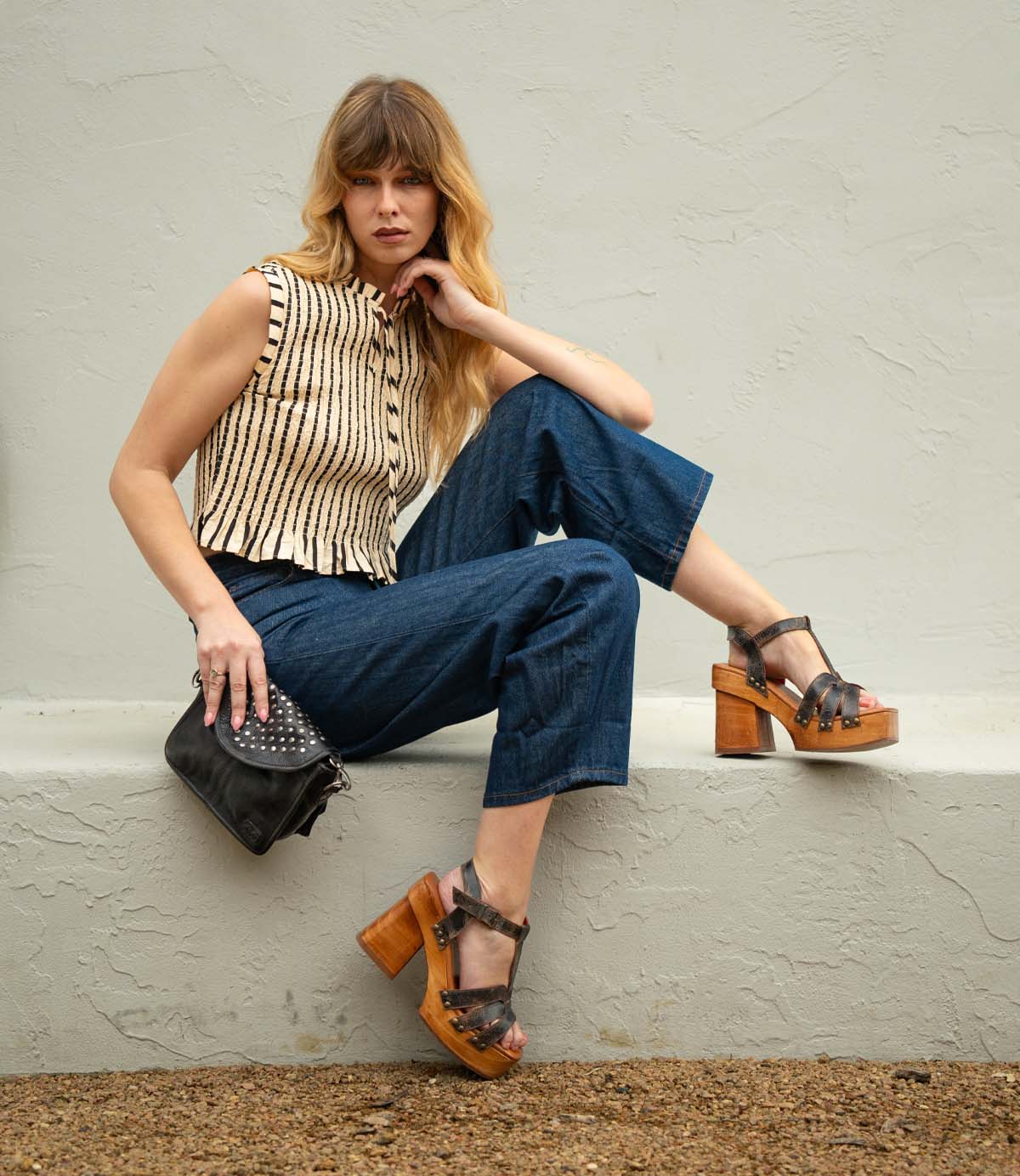 A woman sits on a ledge wearing the Bed Stu Lyxon wooden platform sandals with an open-toe t-strap design, paired with a sleeveless striped top, blue cropped pants, and holding a black studded purse.