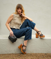 A woman sits on a ledge wearing the Bed Stu Lyxon wooden platform sandals with an open-toe t-strap design, paired with a sleeveless striped top, blue cropped pants, and holding a black studded purse.