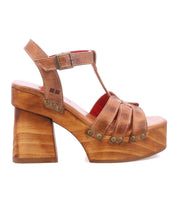 The Bed Stu Lyxon is a brown leather platform sandal featuring an open toe, chunky wooden heel, metal rivets, and a buckled ankle strap, shown in side profile.