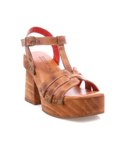 The Lyxon by Bed Stu is a brown leather platform sandal featuring a chunky wooden heel, open-toe T-strap, metal rivet accents, and an adjustable buckle ankle strap.