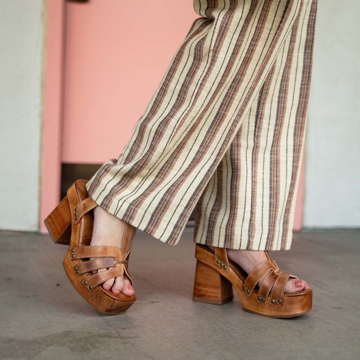 A person stands on a concrete floor in front of a pink door, wearing striped pants and Bed Stu Lyxon brown open-toe platform heel sandals.