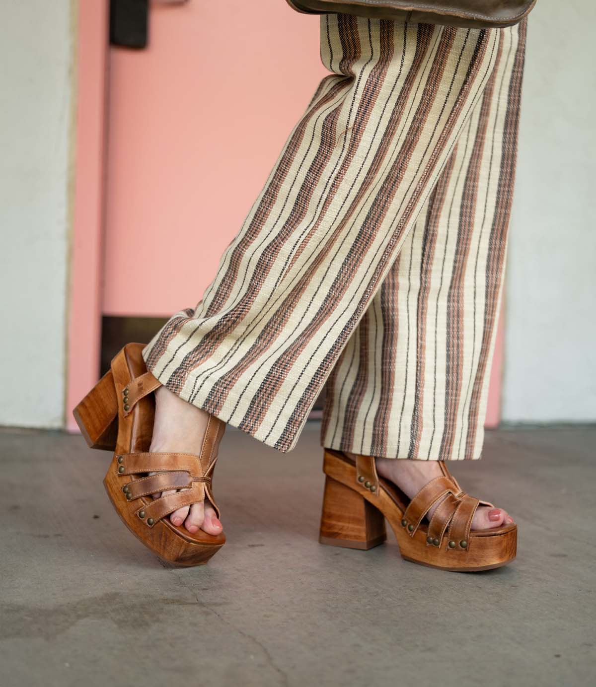 A person stands on a concrete floor in front of a pink door, wearing striped pants and Bed Stu Lyxon brown open-toe platform heel sandals.