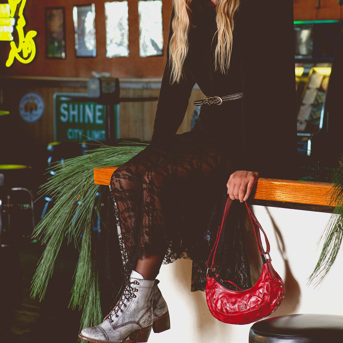 A person in a black lace dress sits on a bar counter, exuding effortless chic with an edgy shoulder bag and gray boots. The Madre handbag by Bed Stu adds a statement-making touch to the scene, complementing the rock 'n' roll edge of the indoor decor.
