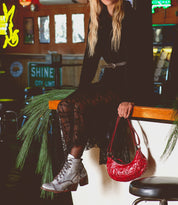 A person in a black lace dress sits on a bar counter, exuding effortless chic with an edgy shoulder bag and gray boots. The Madre handbag by Bed Stu adds a statement-making touch to the scene, complementing the rock 'n' roll edge of the indoor decor.