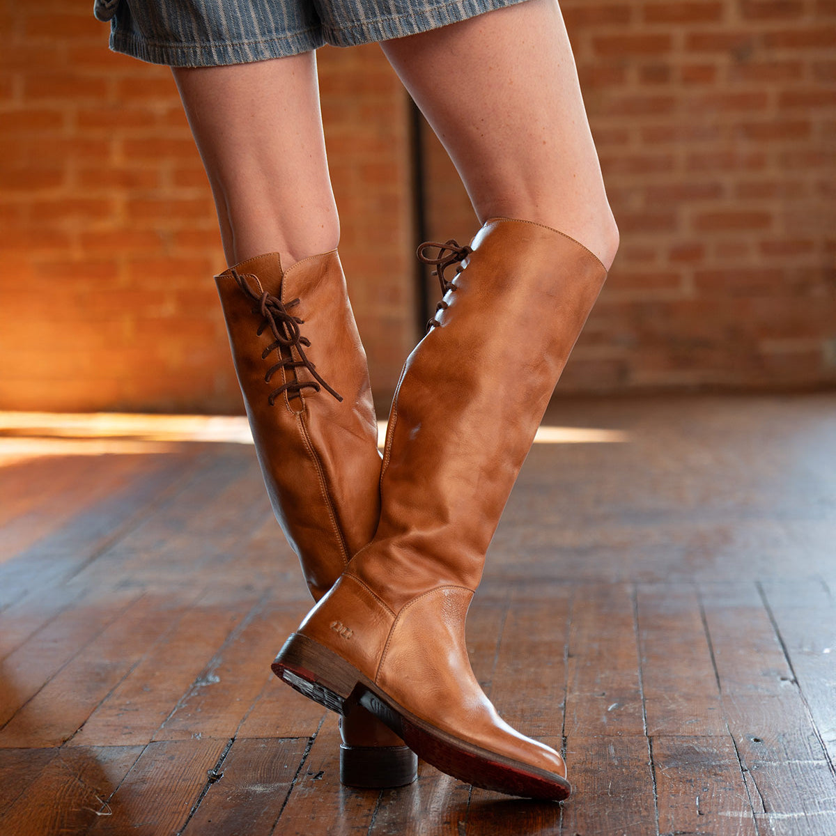 A woman stands on a wooden floor with a brick wall backdrop, wearing Bed Stu Manchester tall tan leather boots with lace-up backs and striped shorts—a stylish twist on an essential women's boot.