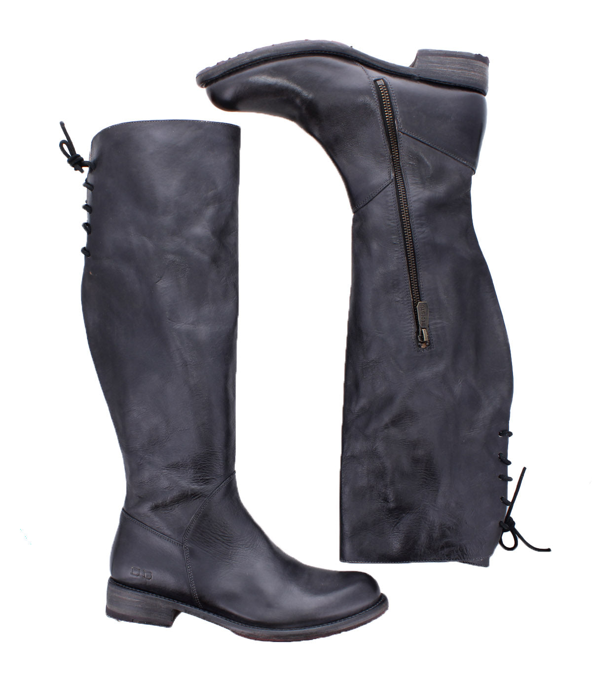 The Bed Stu Manchester boots are black knee-high leather essentials for women, featuring a side zipper and back lace-up detail—one boot stands upright while the other lies on its side.