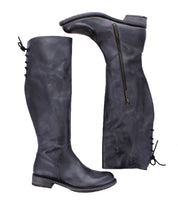 The Bed Stu Manchester boots are black knee-high leather essentials for women, featuring a side zipper and back lace-up detail—one boot stands upright while the other lies on its side.