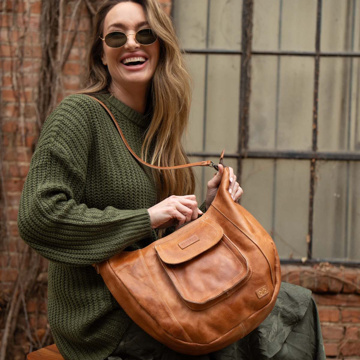 A woman in sunglasses and a green sweater sits smiling by a brick wall and window, holding the Bed Stu Manitoba—a large tan handcrafted bag that's perfect for everyday wear.
