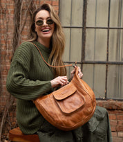 A woman in sunglasses and a green sweater sits smiling by a brick wall and window, holding the Bed Stu Manitoba—a large tan handcrafted bag that's perfect for everyday wear.