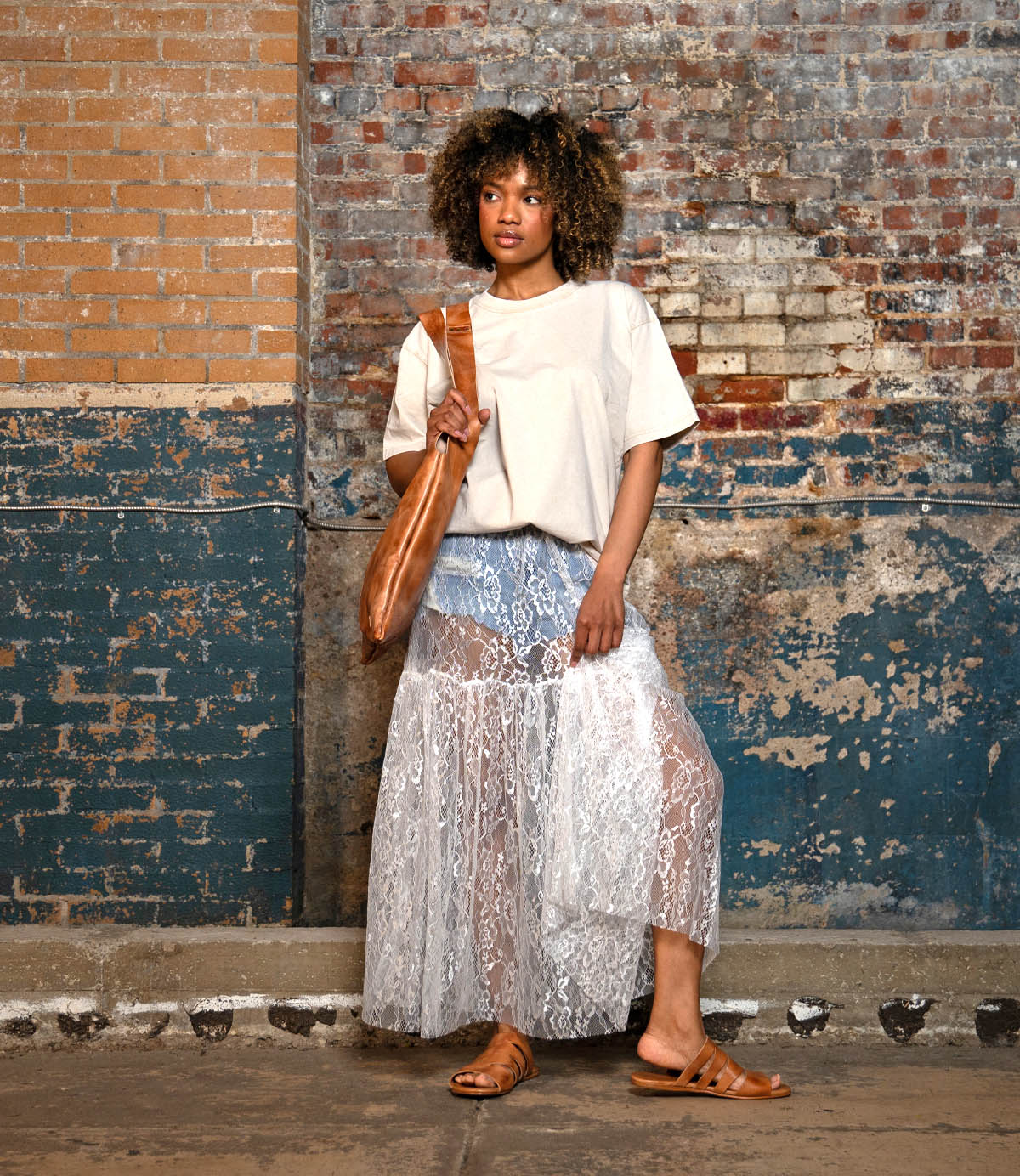A woman stands indoors against a textured brick wall, wearing the Bed Stu Margin slide sandals with leather upper—perfect for effortless summer looks—paired with a white tee, lace skirt over denim shorts, and a tan shoulder bag.