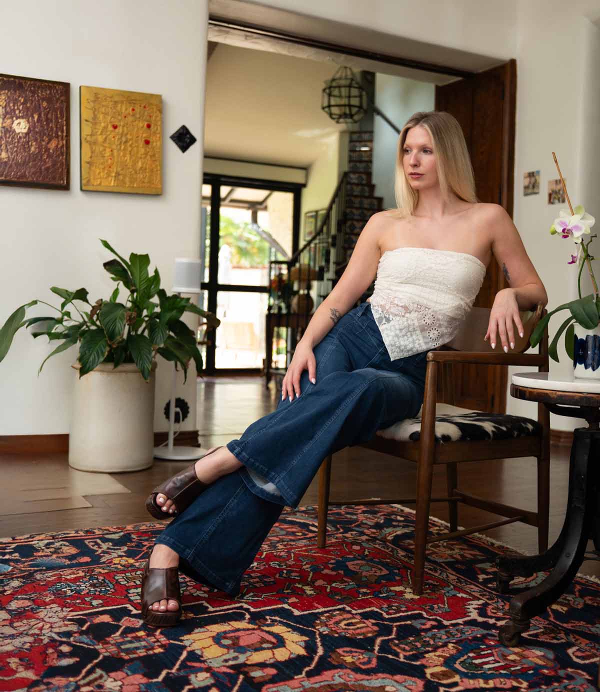 A woman in a white strapless top and blue jeans sits on a wooden chair, wearing Bed Stu Marie heeled clogs made of vegetable tanned leather, in a living room with plants, artwork, and a colorful patterned rug.