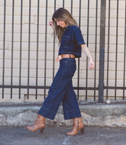 A woman in a full length denim jumpsuit raising up one foot  while walking on cement ground and a iron fence behind her wearing Marjorie boot in tan and a matching Dreamweaver belt by BedStu.