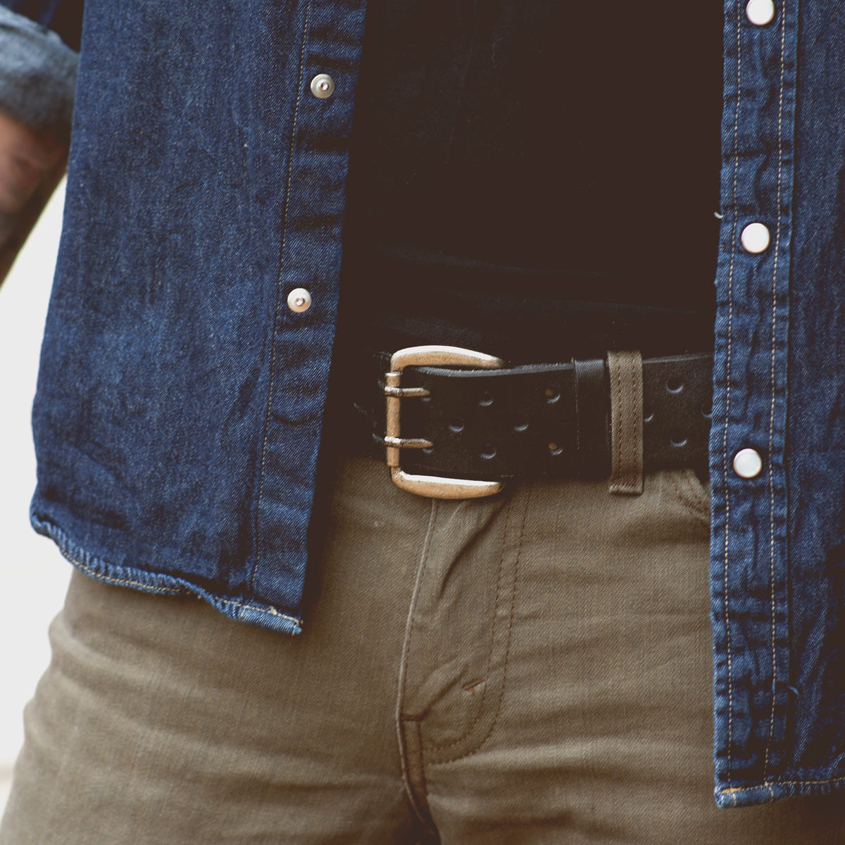 A person wearing a blue denim shirt, olive green pants, and a Bed Stu Mccoy belt with a silver buckle. The shirt is unbuttoned to reveal a black inner shirt, creating a look that seamlessly blends modern aesthetics with timeless design.