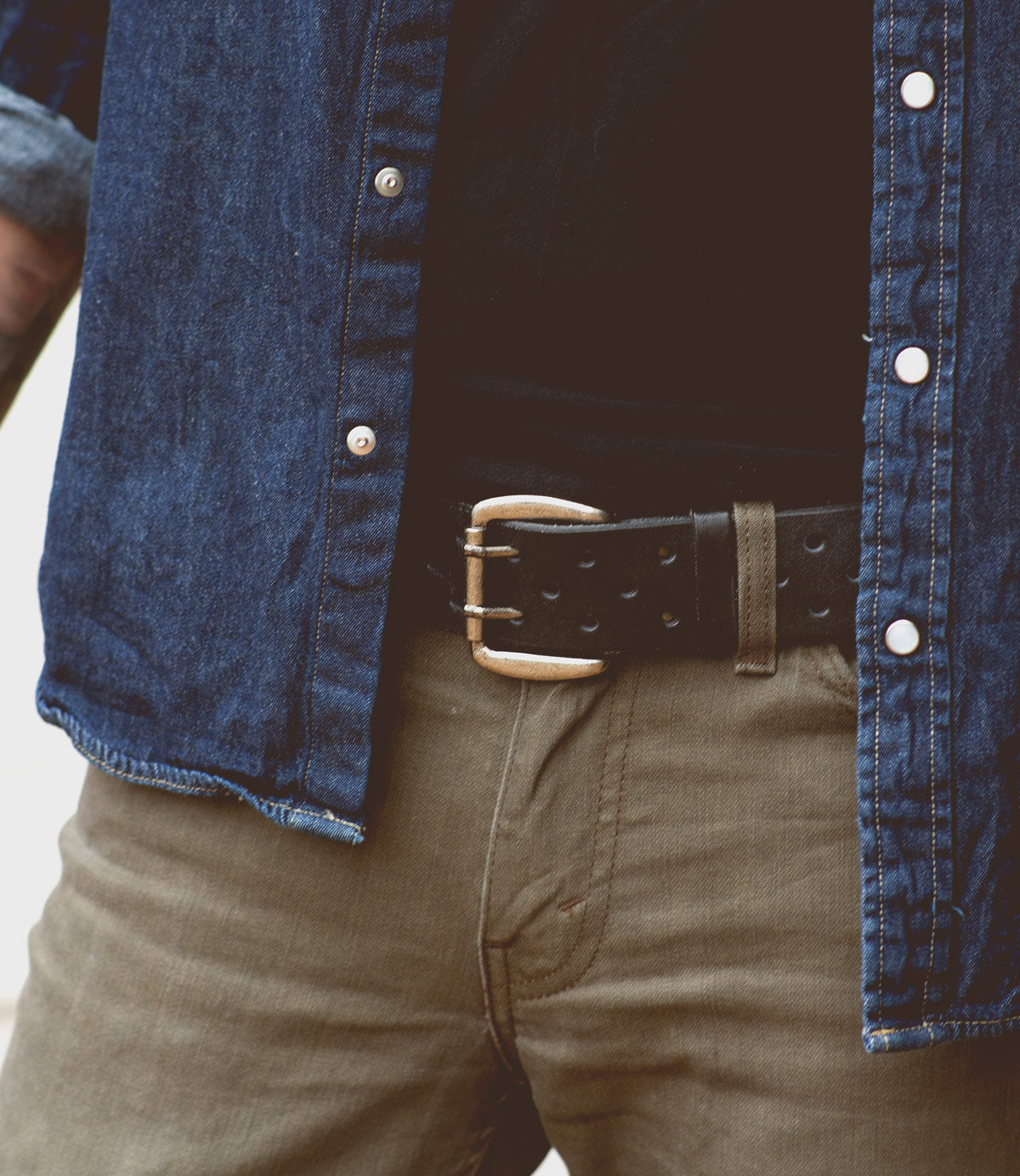 A person wearing a blue denim shirt, olive green pants, and a Bed Stu Mccoy belt with a silver buckle. The shirt is unbuttoned to reveal a black inner shirt, creating a look that seamlessly blends modern aesthetics with timeless design.