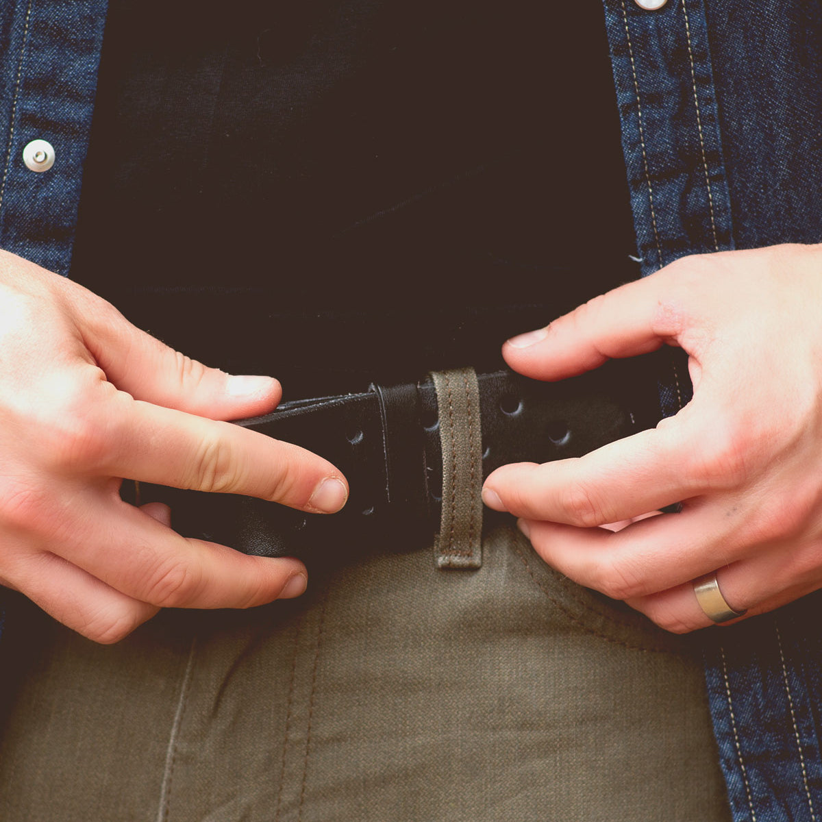 A person is fastening a Bed Stu Mccoy vegetable-tanned leather belt through the belt loops of their beige pants. They are wearing a denim shirt with metal buttons over a black shirt, showcasing a timeless design with modern aesthetics.