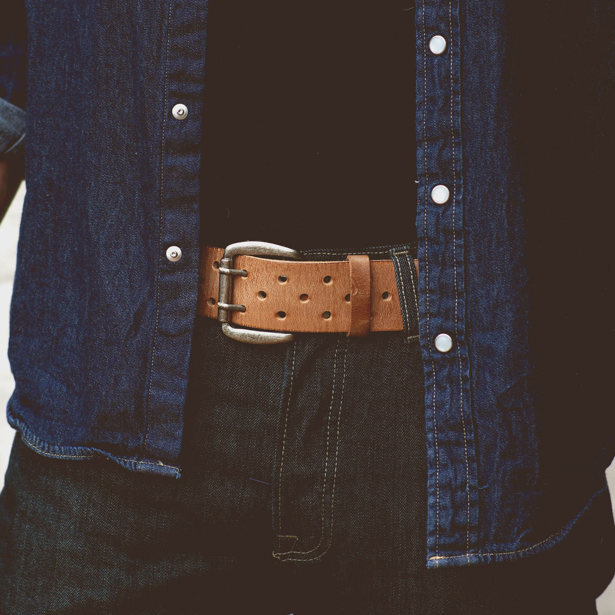 Close-up of a person wearing a denim shirt, black jeans, and a Bed Stu Mccoy brown vegetable-tanned leather belt with a silver buckle.