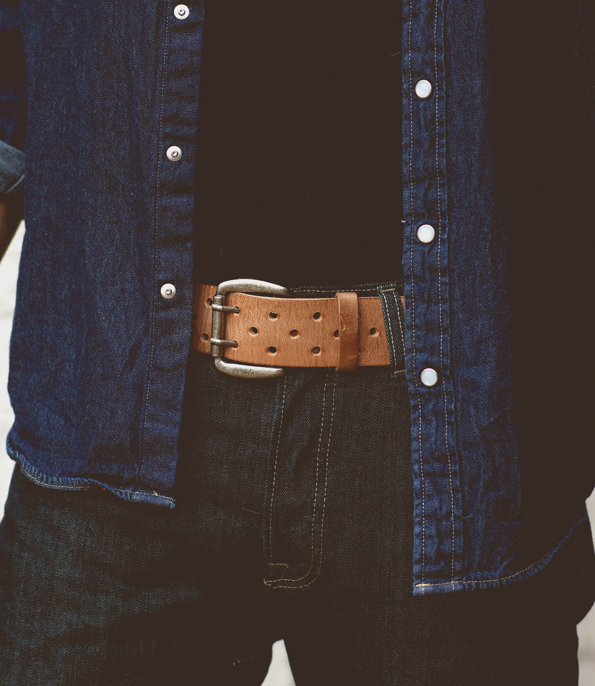 Close-up of a person wearing a denim shirt, black jeans, and a Bed Stu Mccoy brown vegetable-tanned leather belt with a silver buckle.