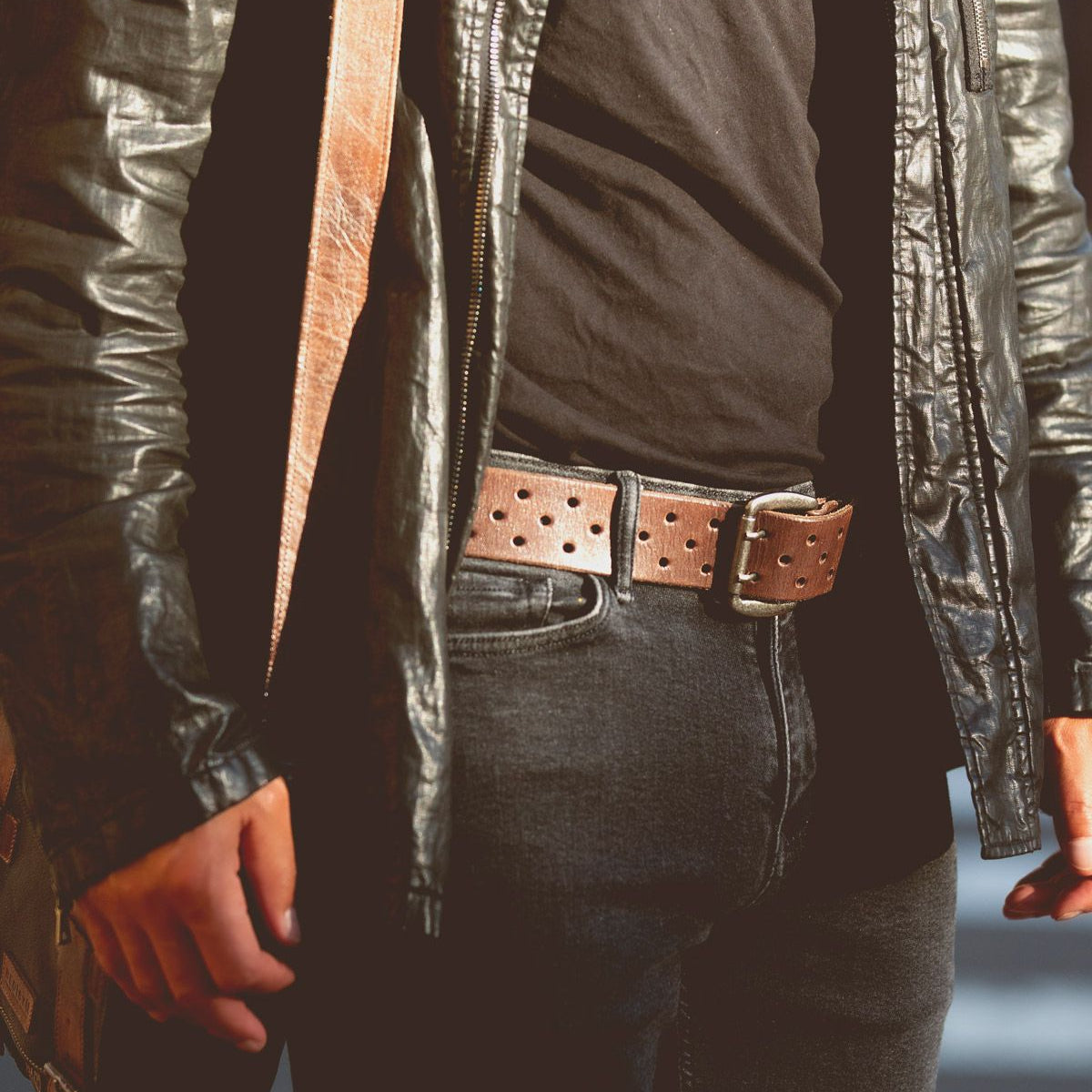 Close-up of a person exuding casual cool style, wearing a black leather jacket, black shirt, dark jeans, and the Proem brown perforated belt with a vintage metal buckle from Bed Stu, standing with arms at their sides.
