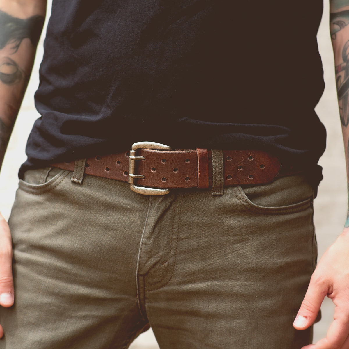 A person wearing a dark t-shirt and olive pants with a timelessly designed brown vegetable-tanned leather belt named Mccoy by Bed Stu. The person's hands are resting near their pockets, partially showing tattoos on their forearms.