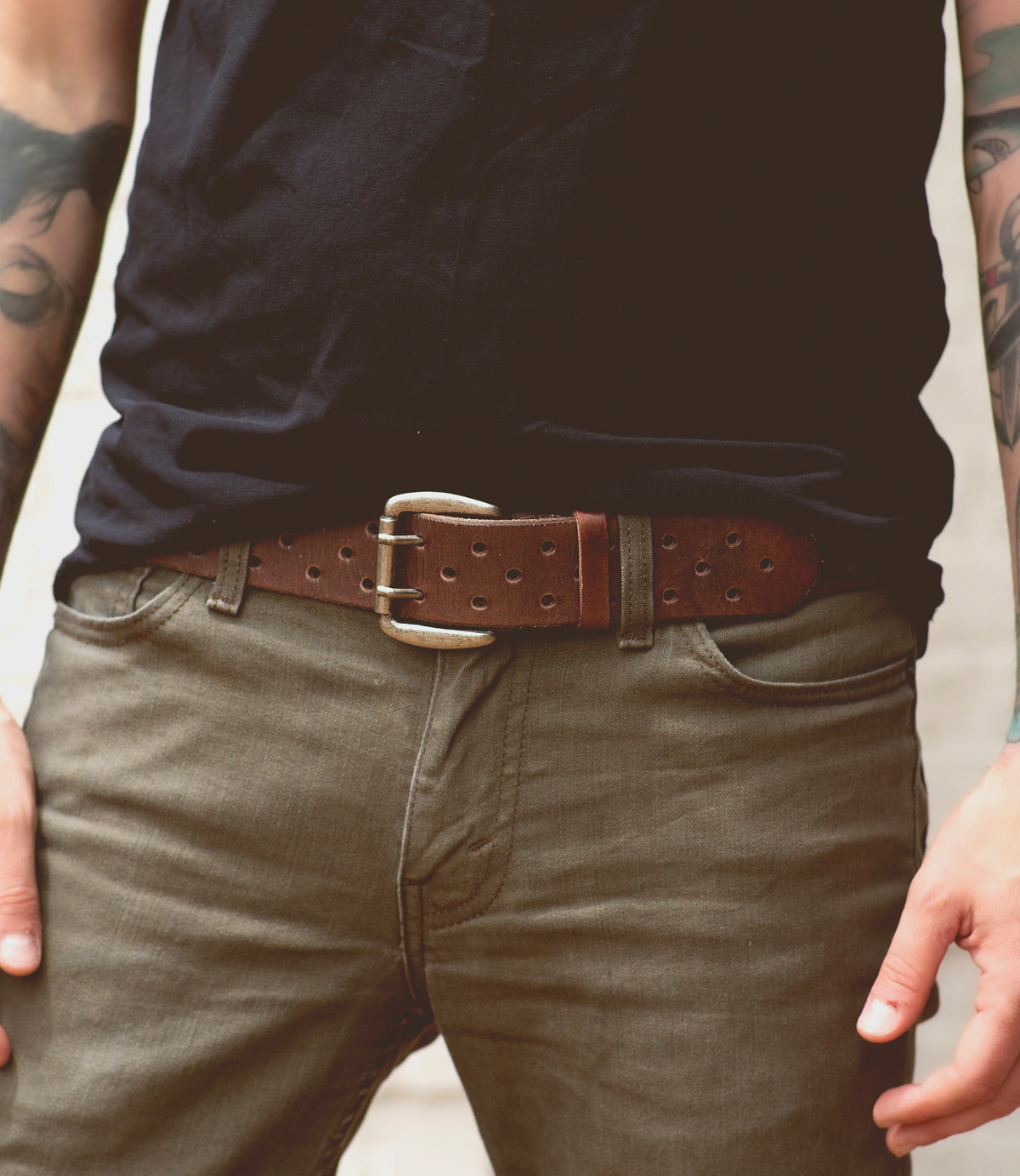 A person wearing a dark t-shirt and olive pants with a timelessly designed brown vegetable-tanned leather belt named Mccoy by Bed Stu. The person's hands are resting near their pockets, partially showing tattoos on their forearms.