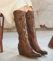 A person wearing white pants is seated and displaying the Meadow cowgirl boots from Bed Stu, which feature decorative flame stitching on their legs. The handcrafted leather boots stand out against the aged, cracked wall and tiled floor.