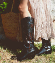 A person stands outdoors on a grassy surface, wearing Bed Stu black cowgirl boots with intricate floral inlay designs and a light-colored flowing skirt, accompanied by a woven straw bag named Meadow.