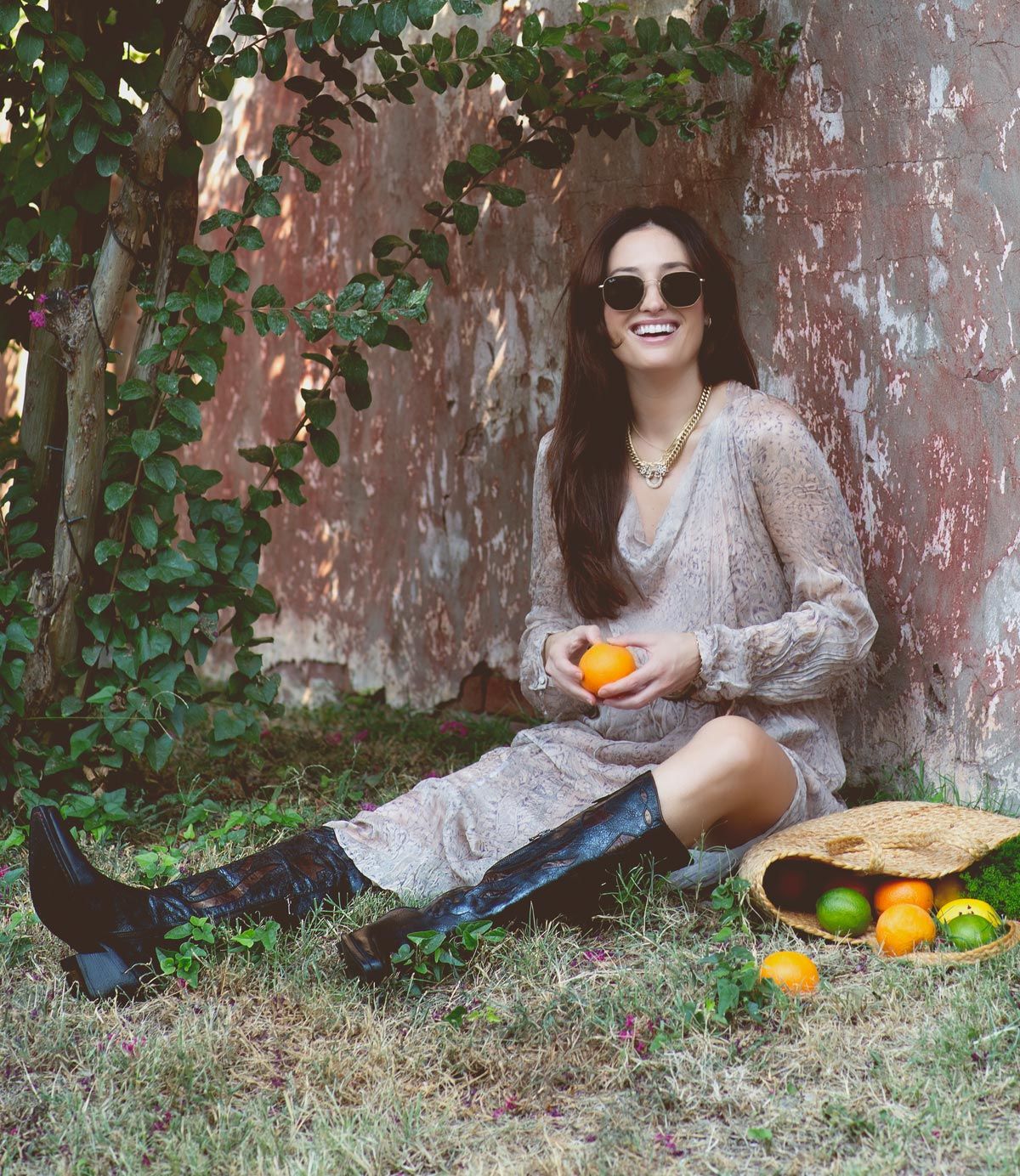 A woman with long hair and sunglasses sits on the ground outdoors, holding an orange. She is wearing a patterned dress and Bed Stu's Meadow cowgirl boots, featuring intricate floral inlay designs. A basket with citrus fruits rests beside her.